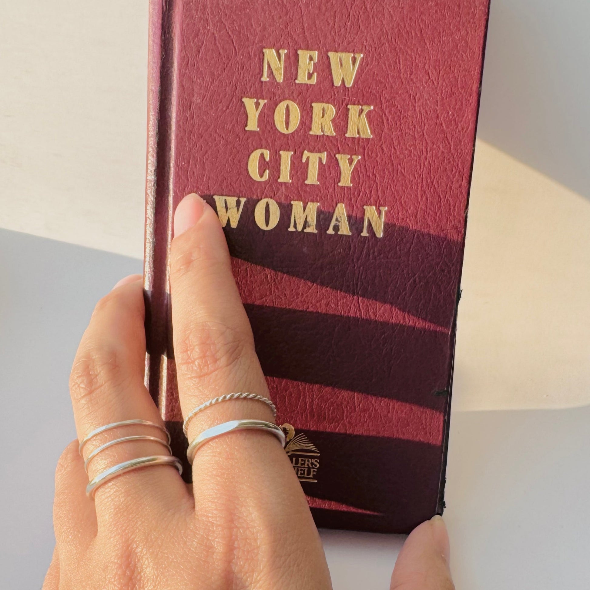 Multiple stacking rings in sterling silver on woman's hand resting against a New York City Woman book